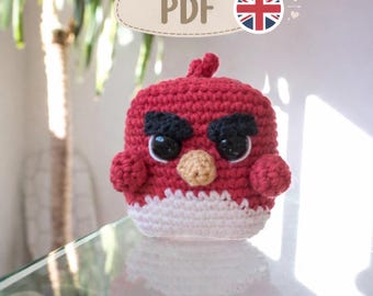 May include: A red, white, and black crocheted bird plush toy with a round body, a small tuft on top, a tan beak, large black eyes, and thick black eyebrows. The text PDF is visible in the upper portion of the image along with a small UK flag icon.