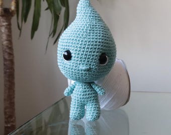 May include: A light blue crochet doll shaped like a teardrop with a pointed top. The doll features two large, round, black plastic safety eyes and a small, simple stitched mouth. The texture is consistent throughout, made of yarn stitches.