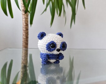 May include: A small, handcrafted crochet panda toy featuring a white head and body with dark blue ears, eye patches, arms, and legs. The panda has large, glossy black safety eyes and a small black nose, with a rounded, compact shape.