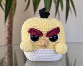 May include: A small, round, yellow crocheted plush toy featuring a white belly, a small black tuft on top, a tan beak, and two large black eyes framed by thick, dark red, angled eyebrows that give it a stern, focused expression.