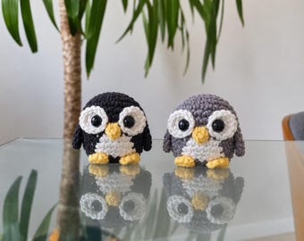 May include: Two small, round, crocheted penguin figures. One is black and white, and the other is gray and white. Both have large black plastic eyes, white eye patches, yellow beaks, and small yellow feet, reflecting on a smooth surface.
