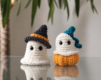 May include: Two small crocheted figures. One is a white ghost wearing a black witch hat with an orange band. The other is a white ghost sitting on an orange pumpkin base, wearing a teal blue hat. Both have black bead eyes and textured yarn.