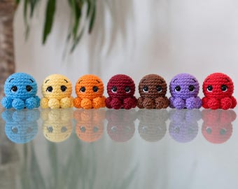 May include: A row of seven small, crocheted octopus toys in blue, yellow, orange, dark red, brown, purple, and red. Each octopus features a round head, multiple small tentacles, and large, glossy black safety eyes with white highlights.