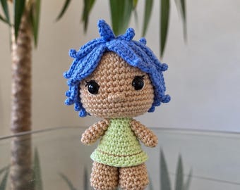 May include: A small crocheted doll featuring a tan yarn body, a light green dress, and bright blue hair styled in short, spiky tufts. The doll has large, round black plastic eyes and a simple stitched nose, crafted with a visible yarn texture.