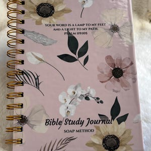 May include: A floral-patterned Bible study journal with a spiral binding. The cover is pale pink and decorated with flower and leaf illustrations. The text on the cover reads "YOUR WORD IS A LAMP TO MY FEET AND A LIGHT TO MY PATH. PSALM 119:105" and "Bible Study Journal SOAP METHOD."