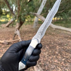 May include: A fixed-blade knife featuring a wavy, patterned Damascus steel blade. The handle is constructed from a translucent blue material with a metallic bolster and pommel, secured by two small brass pins, providing a distinct contrast.