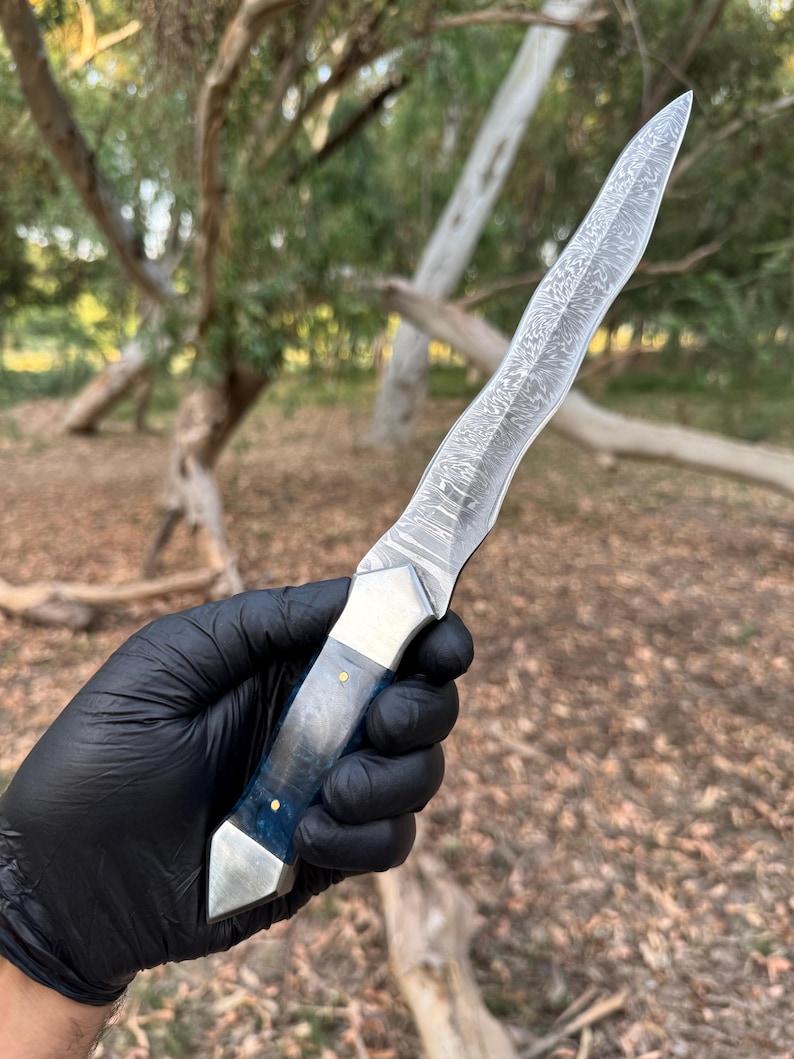 May include: A fixed-blade knife featuring a wavy, patterned Damascus steel blade. The handle is constructed from a translucent blue material with a metallic bolster and pommel, secured by two small brass pins, providing a distinct contrast.