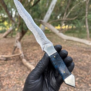 May include: A fixed-blade knife featuring a wavy, patterned Damascus steel blade, a polished metal bolster, and a dark blue, marbled resin handle secured with two small brass pins, finished with a pointed metal pommel at the base of the grip.