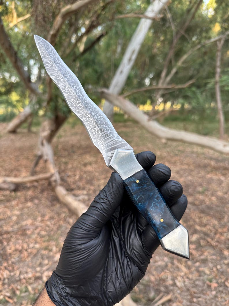 May include: A fixed-blade knife featuring a wavy, patterned Damascus steel blade, a polished metal bolster, and a dark blue, marbled resin handle secured with two small brass pins, finished with a pointed metal pommel at the base of the grip.