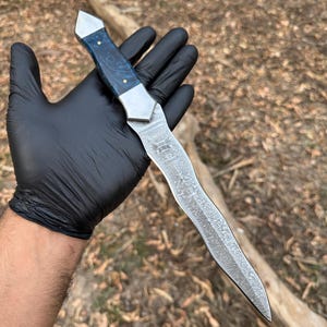 May include: A fixed-blade knife featuring a wavy, patterned Damascus steel blade and a handle made of polished blue resin with metallic accents. The handle is secured with two small brass pins, and the blade has a distinct, flowing, organic shape.