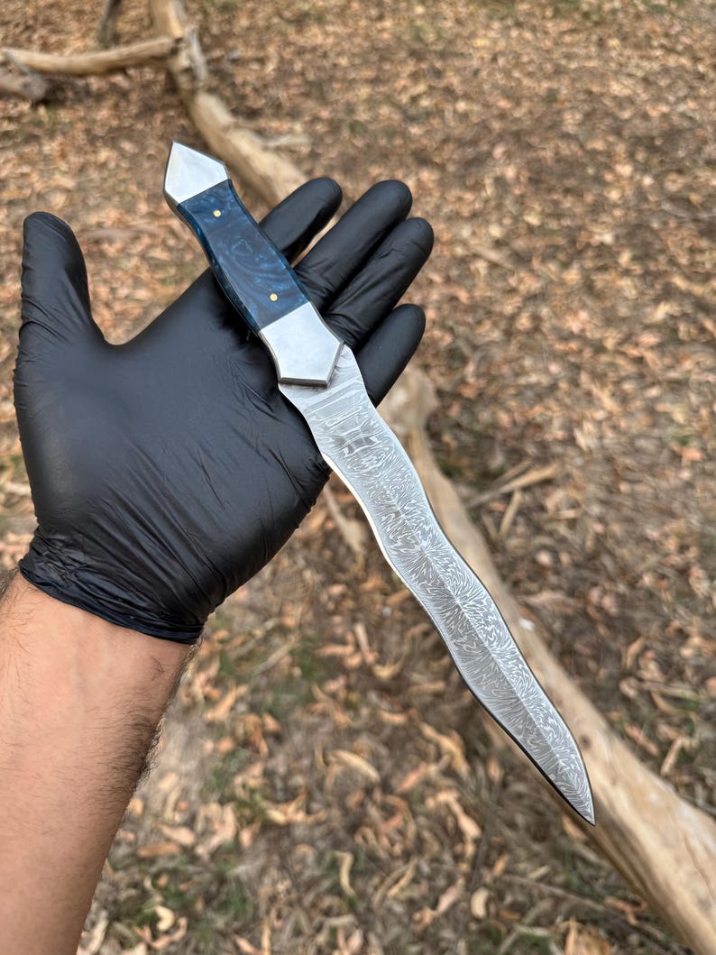 May include: A fixed-blade knife featuring a wavy, patterned Damascus steel blade and a handle made of polished blue resin with metallic accents. The handle is secured with two small brass pins, and the blade has a distinct, flowing, organic shape.