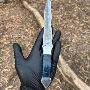 May include: A fixed-blade knife featuring a wavy, patterned Damascus steel blade and a handle crafted from blue marbled resin with two small brass pins, finished with a polished metal bolster and a pointed metal pommel at the base.