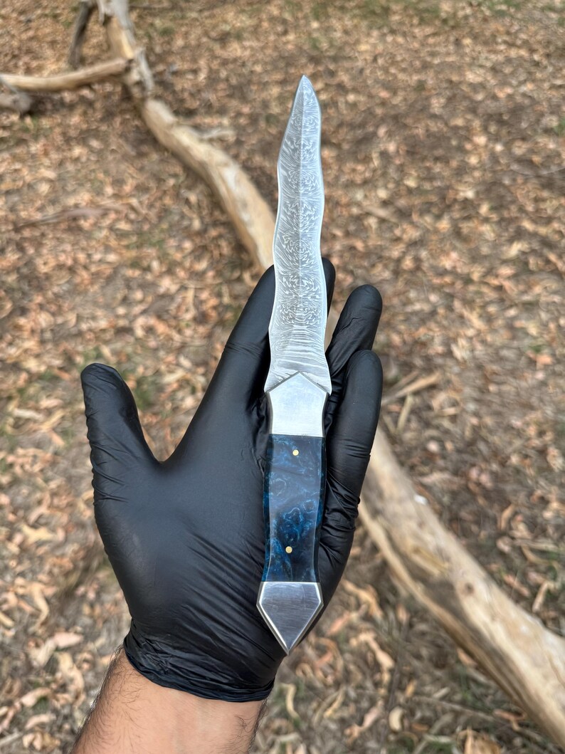May include: A fixed-blade knife featuring a wavy, patterned Damascus steel blade and a handle crafted from blue marbled resin with two small brass pins, finished with a polished metal bolster and a pointed metal pommel at the base.