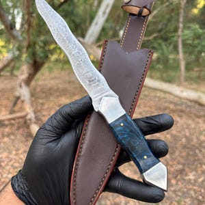 May include: A fixed-blade knife featuring a wavy Damascus steel blade, a polished metal bolster, and a deep blue marbled resin handle with two brass pins, accompanied by a brown leather sheath with contrast stitching and a snap button closure.