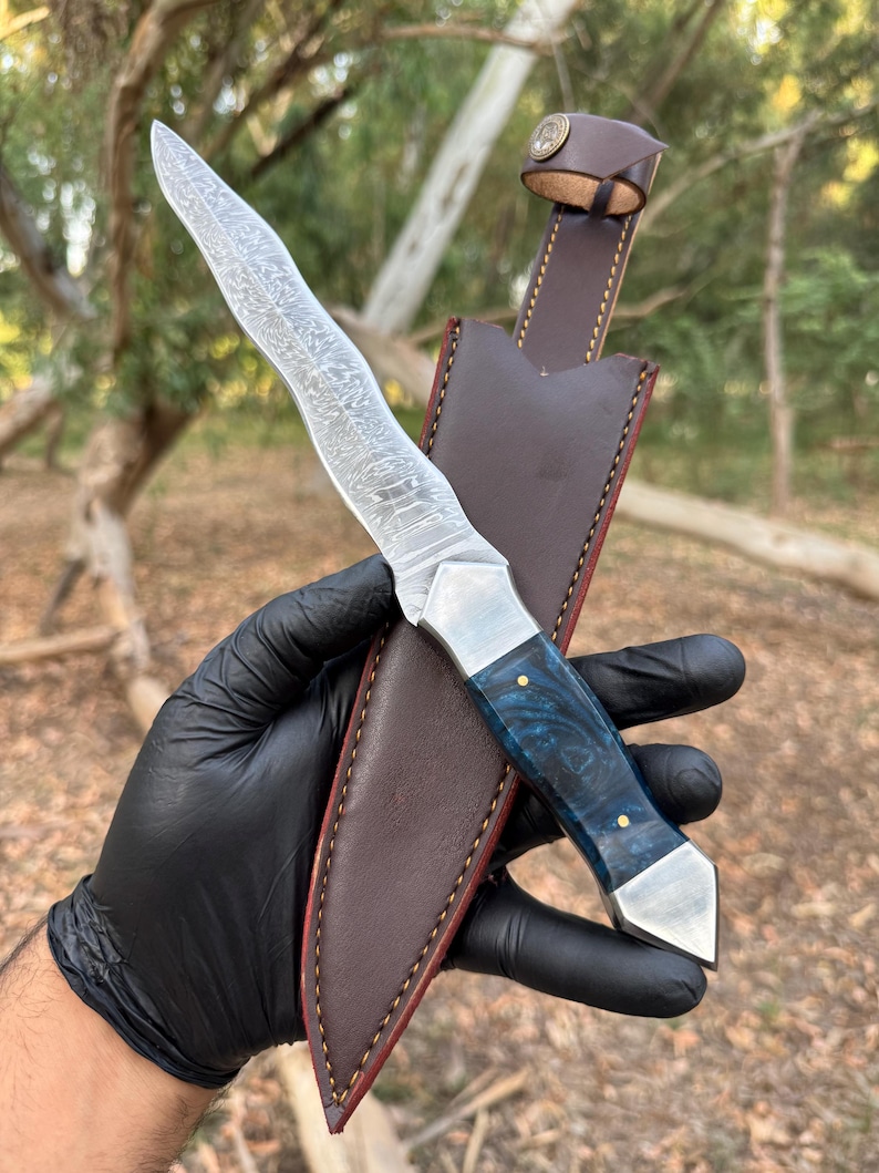 May include: A fixed-blade knife featuring a wavy Damascus steel blade, a polished metal bolster, and a deep blue marbled resin handle with two brass pins, accompanied by a brown leather sheath with contrast stitching and a snap button closure.