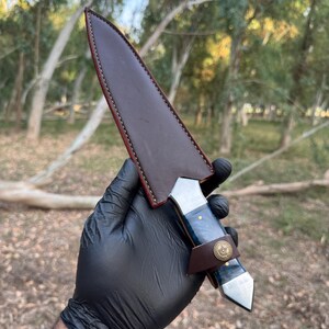 May include: A fixed-blade knife with a dark blue marbled handle and a metallic pommel, secured in a dark brown leather sheath. The sheath features visible stitching along the edges and a small leather strap with a circular brass-colored button.