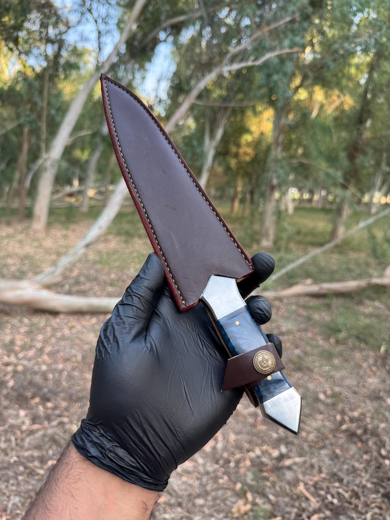 May include: A fixed-blade knife with a dark blue marbled handle and a metallic pommel, secured in a dark brown leather sheath. The sheath features visible stitching along the edges and a small leather strap with a circular brass-colored button.