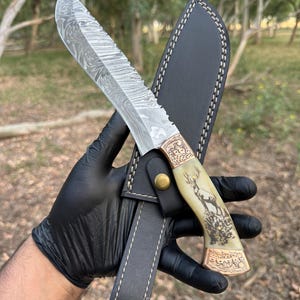 May include: A Damascus steel hunting knife with a decorative handle featuring a deer illustration and a black leather sheath. The blade has a unique pattern. The handle is cream-colored with gold-colored accents.