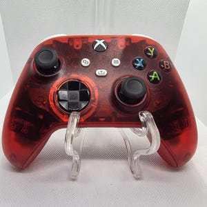 Może przedstawiać: Przezroczysty czerwony kontroler Xbox z czarnymi gałkami i przyciskami. Kontroler ma białą górę i jest wyświetlany na przezroczystym stojaku. Przyciski funkcyjne są w kolorze zielonym, niebieskim, żółtym i czerwonym.