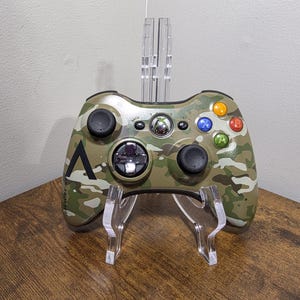 May include: A camouflage patterned video game controller featuring a green, brown, and white colour scheme. The device includes two black analogue sticks, a directional pad, a central Xbox guide button, and four coloured action buttons labelled A, B, X, and Y.