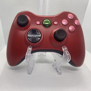 May include: A matte red video game controller featuring two black analog sticks, a black directional pad, and four light pink action buttons. The center includes a metallic green circular button, along with small pink start and back buttons.