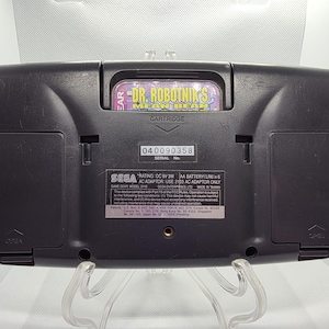 K&ouml;nnte beinhalten: Schwarze Sega Game Gear-Handheld-Konsole mit eingelegter Spielkassette. Die Kassette ist mit "Dr. Robotnik's Mean Bean" beschriftet. Die R&uuml;ckseite der Konsole hat eine Seriennummer und weitere gedruckte Informationen.