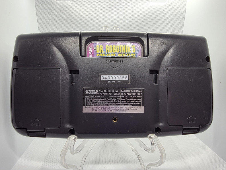 K&ouml;nnte beinhalten: Schwarze Sega Game Gear-Handheld-Konsole mit eingelegter Spielkassette. Die Kassette ist mit "Dr. Robotnik's Mean Bean" beschriftet. Die R&uuml;ckseite der Konsole hat eine Seriennummer und weitere gedruckte Informationen.