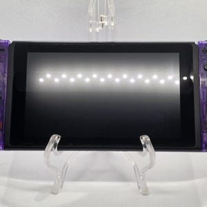 Peut inclure: Une console Nintendo Switch violette avec des manettes Joy-Con violet transparentes. La console a un écran noir et est présentée sur un support en plastique transparent. Les boutons et les composants internes sont visibles.