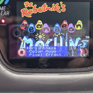 K&ouml;nnte beinhalten: Schwarze Sega Game Gear-Handheld-Konsole mit dem Spiel "Dr. Robotnik's Mean Bean Machine". Der Bildschirm zeigt den Titel des Spiels und Men&uuml;optionen, einschlie&szlig;lich Helligkeits- und Farbmoduseinstellungen. Die Betriebsanzeige der Konsole leuchtet.