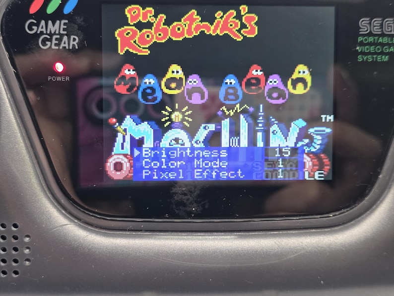 K&ouml;nnte beinhalten: Schwarze Sega Game Gear-Handheld-Konsole mit dem Spiel "Dr. Robotnik's Mean Bean Machine". Der Bildschirm zeigt den Titel des Spiels und Men&uuml;optionen, einschlie&szlig;lich Helligkeits- und Farbmoduseinstellungen. Die Betriebsanzeige der Konsole leuchtet.