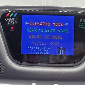 K&ouml;nnte beinhalten: Nahaufnahme einer schwarzen Sega Game Gear Handheld-Videospielkonsole. Der Bildschirm zeigt ein blaues Men&uuml; mit Optionen wie "Scenario Mode" und "Gear to Gear Mode". Die Konsole hat einen Power-Knopf und der Text "Game Gear" ist sichtbar.