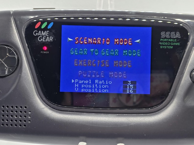 K&ouml;nnte beinhalten: Nahaufnahme einer schwarzen Sega Game Gear Handheld-Videospielkonsole. Der Bildschirm zeigt ein blaues Men&uuml; mit Optionen wie "Scenario Mode" und "Gear to Gear Mode". Die Konsole hat einen Power-Knopf und der Text "Game Gear" ist sichtbar.