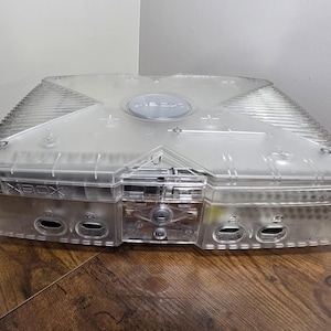 以下が含まれることがあります： 透明なプラスチック製のXboxビデオゲーム機。上部には特徴的なX字型のデザインが施され、中央にはXboxのロゴがあります。前面パネルには、4つのコントローラーポート、電源ボタン、イジェクトボタンが配置されており、ケース越しに内部構造が見えます。