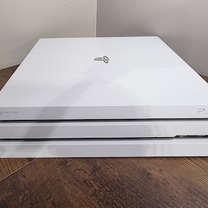 Puede incluir: Una consola de videojuegos Sony PlayStation 4 Pro de color blanco con un diseño rectangular elegante de varios niveles. La superficie superior muestra el logotipo de PlayStation, mientras que el panel frontal incluye la marca Sony y el logo PS4.