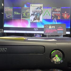 May include: A black Xbox 360 game console with a glowing green power button, positioned in front of a monitor displaying a digital game menu with the title Alpha Protocol and various other game icons, alongside a small rectangular business card.