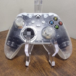 May include: A transparent wireless video game controller featuring a clear outer shell, a black directional pad, two clear analog sticks, and four colored action buttons labeled X, Y, A, and B. The controller includes a central Xbox logo button.