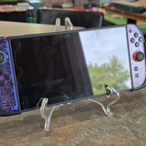 May include: A black Nintendo Switch console with purple and blue patterned Joy-Con controllers. The console is displayed on a clear plastic stand, showcasing the screen. The controllers feature a circuit board design with purple buttons and a black grip.