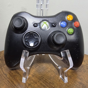 Op de afbeelding: Een zwarte draadloze videogamecontroller met twee analoge sticks, een richtingspad en vier gekleurde actieknoppen met de labels A, B, X en Y. Het midden bevat een ronde Xbox-guideknop, samen met kleine Back- en Start-knoppen.