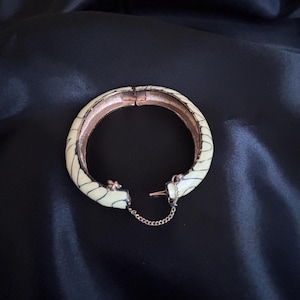 Pode incluir: Pulseira de bracelete articulada creme com detalhes lineares pretos. A pulseira tem um interior dourado e uma corrente de segurança. O fecho está aberto, revelando o interior. A pulseira está em um fundo preto.