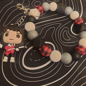 Könnte beinhalten: Ein Schlüsselanhänger mit Perlen und einem Cartoon-Figur-Anhänger. Das Armband hat graue, schwarze und rot-schwarz karierte Perlen. Der Anhänger ist eine Cartoonfigur mit einer roten Gitarre.