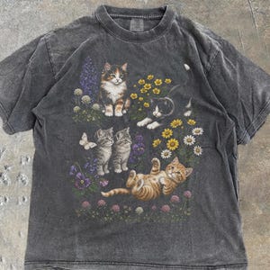 Könnte beinhalten: Ein anthrazitfarbenes Kurzarm-T-Shirt mit einem Grafikdruck von vier Katzen in einer Gartenumgebung. Das Design zeigt eine Glückskatze, eine schwarz-weiße Katze, zwei graue getigerte Kätzchen und ein orangefarbenes getigertes Kätzchen inmitten von Blumen.