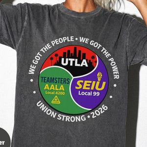 Op de afbeelding: Donkergrijs t-shirt met een cirkelvormige afbeelding. De afbeelding bevat de tekst "WE GOT THE PEOPLE", "WE GOT THE POWER", "UTLA", "TEAMSTERS AALA" en "SEIU Local 99". Het shirt heeft ook de tekst "UNION STRONG 2026".