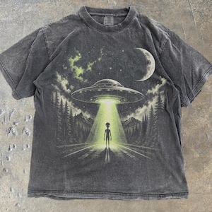 Könnte beinhalten: Ein anthrazitfarbenes Kurzarm-T-Shirt mit einem Grafikdruck, der eine fliegende Untertasse über einer Waldstraße zeigt. Ein heller grüner Lichtstrahl scheint vom Raumschiff auf eine Alien-Figur herab. Ein Halbmond und Sterne sind zu sehen.
