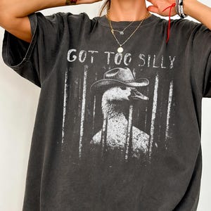 以下が含まれることがあります： 「GOT TOO SILLY」の文字と、カウボーイハットをかぶったアヒルのグラフィックが描かれたダークグレーのTシャツ。白いデザインがヴィンテージ感とダメージ感を演出しています。