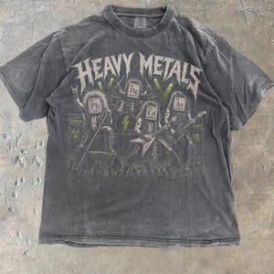May include: A charcoal gray short-sleeve t-shirt featuring a graphic of a band with periodic table element symbols as heads. The text HEAVY METALS is printed above the band members, who are playing instruments on a stage with green accents.