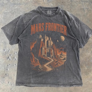 May include: A charcoal gray short-sleeve t-shirt featuring a graphic print in orange and black. The design depicts a castle on a rocky landscape with the text MARS FRONTIER arched above. Two planets are visible in the sky above the structure.