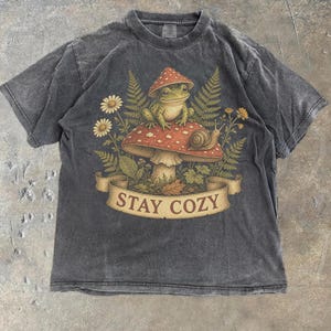 Op de afbeelding: Een antracietgrijs t-shirt met korte mouwen en een grafische print van een groene kikker die op een grote rode paddenstoel met witte stippen zit. Een kleine slak rust op de hoed, omringd door varens en madeliefjes, met een banner met STAY COZY.