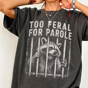 May include: Dark gray t-shirt with the text "TOO FERAL FOR PAROLE" above a graphic of a raccoon behind bars. The raccoon is in black and white, with its hands on the bars. The shirt has a vintage, worn look.