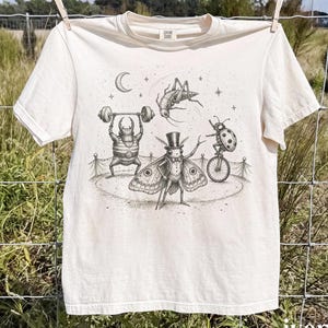 May include: An ivory short-sleeved t-shirt featuring a black ink illustration of insects performing in a circus. The graphic depicts a beetle lifting weights, a moth in a top hat, a ladybug on a unicycle, and a grasshopper jumping under a crescent moon.