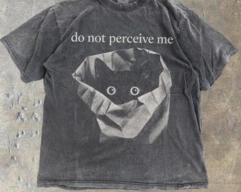 Peut inclure: Un t-shirt à manches courtes gris anthracite avec un aspect délavé. L'avant présente un graphique d'un chat noir sortant d'un sac en papier, avec le texte blanc "do not perceive me" imprimé au-dessus de l'illustration du chat.